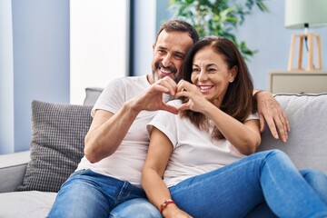Middle age man and woman couple doing heart gesture with hands sitting on sofa at home