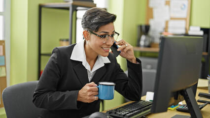 Young beautiful hispanic woman business worker talking on smartphone drinking coffee working at office