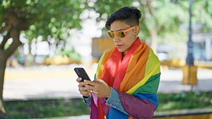 Young beautiful hispanic woman using smartphone wearing rainbow flag at park