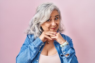 Middle age woman with grey hair standing over pink background laughing nervous and excited with...