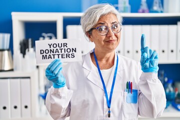 Middle age woman with grey hair working at scientist laboratory holding your donation matters banner surprised with an idea or question pointing finger with happy face, number one