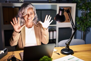 Middle age woman with grey hair working using computer laptop late at night showing and pointing up...
