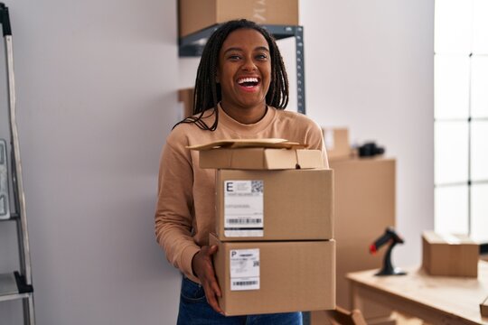 Young african american with braids working at small business ecommerce holding packages smiling and laughing hard out loud because funny crazy joke.