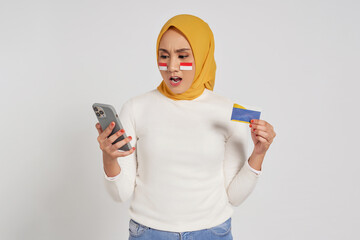Shocked young Asian Muslim woman wearing a hijab holding smartphone and credit card isolated over...
