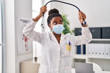 Naklejka premium African american woman wearing doctor uniform and medical mask holding stethoscope at clinic
