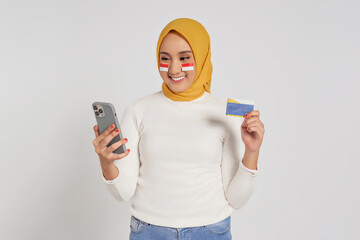 Smiling young Asian Muslim woman wearing a hijab holding smartphone and credit card isolated over...