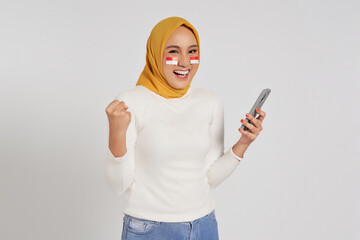Excited young Asian Muslim woman wearing a hijab using smartphone and celebrating Indonesian...