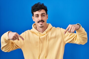 Hispanic man standing over blue background looking confident with smile on face, pointing oneself...