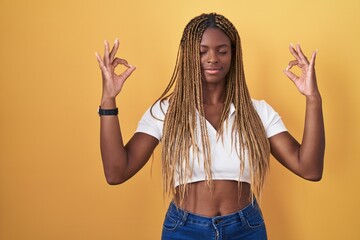 African american woman with braided hair standing over yellow background relaxed and smiling with eyes closed doing meditation gesture with fingers. yoga concept.