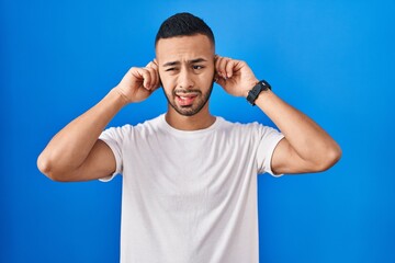 Young hispanic man standing over blue background covering ears with fingers with annoyed expression for the noise of loud music. deaf concept.
