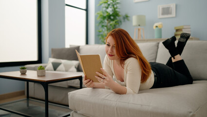 Young redhead woman reading book lying on sofa at home