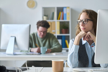 Young businesswoman in eyeglasses sitting at her workplace with computer with her colleague working in background