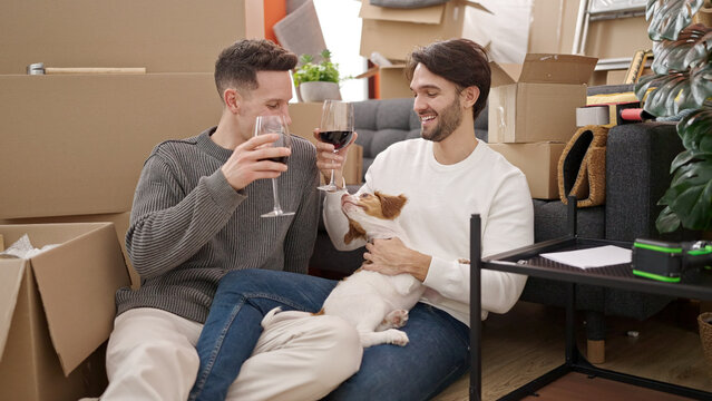 Two men couple drinking glass of wine sitting on floor with dog at new home