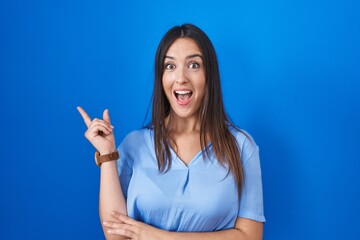 Fototapeta premium Young brunette woman standing over blue background with a big smile on face, pointing with hand finger to the side looking at the camera.
