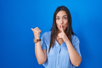 Young brunette woman standing over blue background asking to be quiet with finger on lips pointing with hand to the side. silence and secret concept.