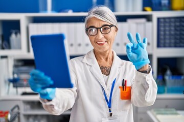 Middle age woman with grey hair working at scientist laboratory doing video call doing ok sign with fingers, smiling friendly gesturing excellent symbol