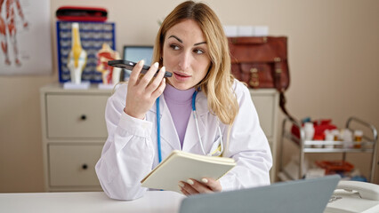Young beautiful hispanic woman doctor sending voice message by smartphone reading notebook at clinic
