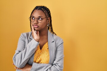 African american woman with braids standing over yellow background with hand on chin thinking about question, pensive expression. smiling with thoughtful face. doubt concept.