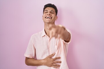 Young hispanic man standing over pink background laughing at you, pointing finger to the camera with hand over body, shame expression