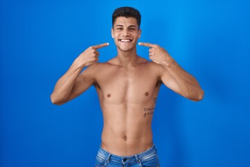 Young hispanic man standing shirtless over blue background smiling cheerful showing and pointing with fingers teeth and mouth. dental health concept.