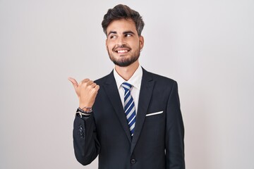 Young hispanic man with tattoos wearing business suit and tie smiling with happy face looking and pointing to the side with thumb up.