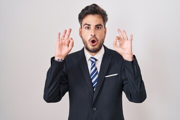 Young hispanic man with tattoos wearing business suit and tie looking surprised and shocked doing ok approval symbol with fingers. crazy expression
