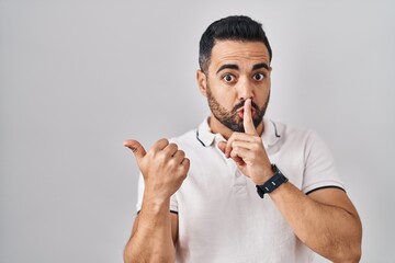 Young hispanic man with beard wearing casual clothes over white background asking to be quiet with finger on lips pointing with hand to the side. silence and secret concept.