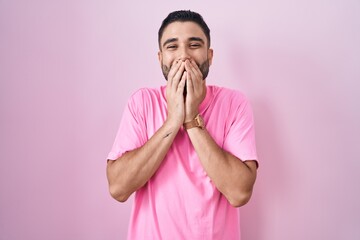 Hispanic young man standing over pink background laughing and embarrassed giggle covering mouth with hands, gossip and scandal concept