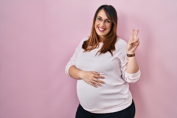 Pregnant woman standing over pink background smiling with happy face winking at the camera doing...