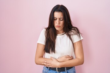 Young brunette woman standing over pink background with hand on stomach because indigestion,...