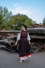 А woman in Ukrainian national embroidered dress vyshyvanka near destroyed russian tank and cars. - 619777873