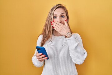 Young blonde woman using smartphone typing message shocked covering mouth with hands for mistake. secret concept.