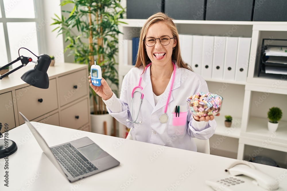 Poster young blonde doctor woman holding glucometer and sweets sticking tongue out happy with funny express