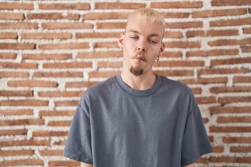 Young caucasian man standing over bricks wall making fish face with lips, crazy and comical gesture. funny expression.