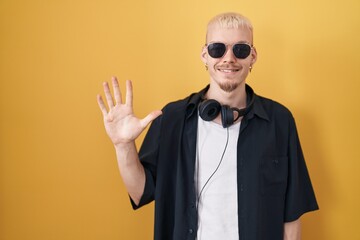Young caucasian man wearing sunglasses standing over yellow background showing and pointing up with fingers number five while smiling confident and happy.