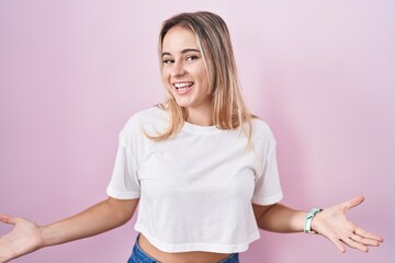 Young blonde woman standing over pink background smiling cheerful with open arms as friendly welcome, positive and confident greetings