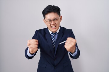 Young asian man wearing business suit and tie angry and mad raising fists frustrated and furious...