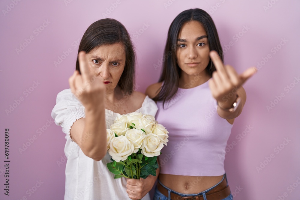 Poster Hispanic mother and daughter holding bouquet of white flowers showing middle finger, impolite and rude fuck off expression