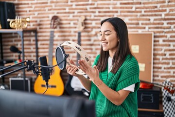 Obraz na płótnie Canvas Young hispanic woman musician playing tambourine at music studio