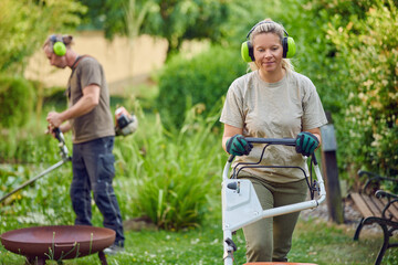 Young Female gardener using a lawn mower while her colleague or bosses is trimming the garden in the background