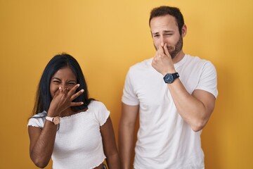 Interracial couple standing over yellow background smelling something stinky and disgusting,...