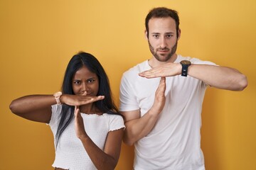 Interracial couple standing over yellow background doing time out gesture with hands, frustrated...