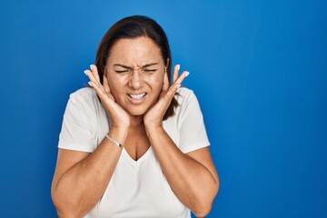Hispanic mature woman standing over blue background covering ears with fingers with annoyed expression for the noise of loud music. deaf concept.