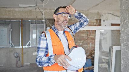 Middle age man builder holding hardhat sweating at construction site