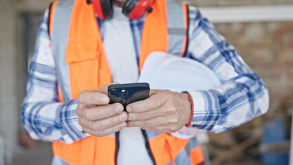 Middle age man builder holding hardhat using smartphone at construction site