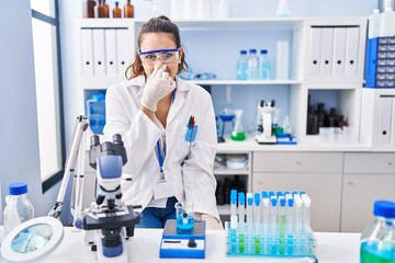 Fototapeta premium Young hispanic woman working at scientist laboratory smelling something stinky and disgusting, intolerable smell, holding breath with fingers on nose. bad smell