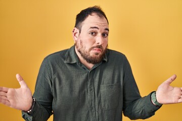 Plus size hispanic man with beard standing over yellow background clueless and confused expression...