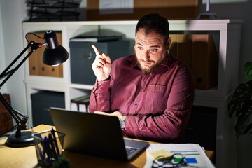 Plus size hispanic man with beard working at the office at night with a big smile on face, pointing with hand and finger to the side looking at the camera.