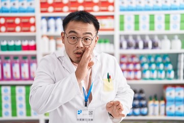 Chinese young man working at pharmacy drugstore hand on mouth telling secret rumor, whispering malicious talk conversation