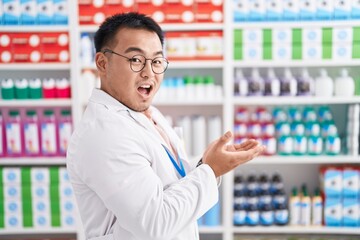 Chinese young man working at pharmacy drugstore pointing aside with hands open palms showing copy space, presenting advertisement smiling excited happy